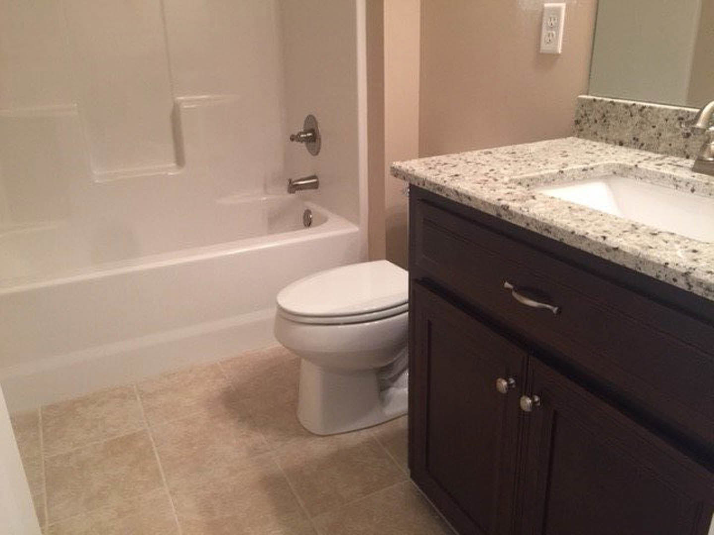 White porcelain sink with chrome faucet set in a light stone countertop, adjacent to a modern toilet, neutral tile flooring, and a white wall outlet visible nearby.