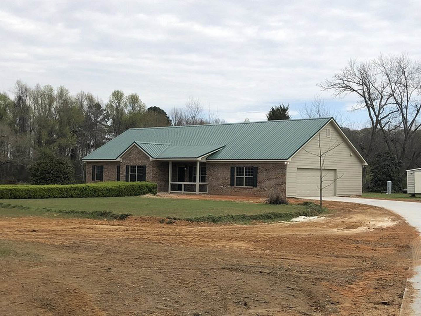 Green metal roof atop a brick house with white-framed windows, covered porch with exterior light, grassy dirt field and hedge in foreground, cloudy sky overhead.