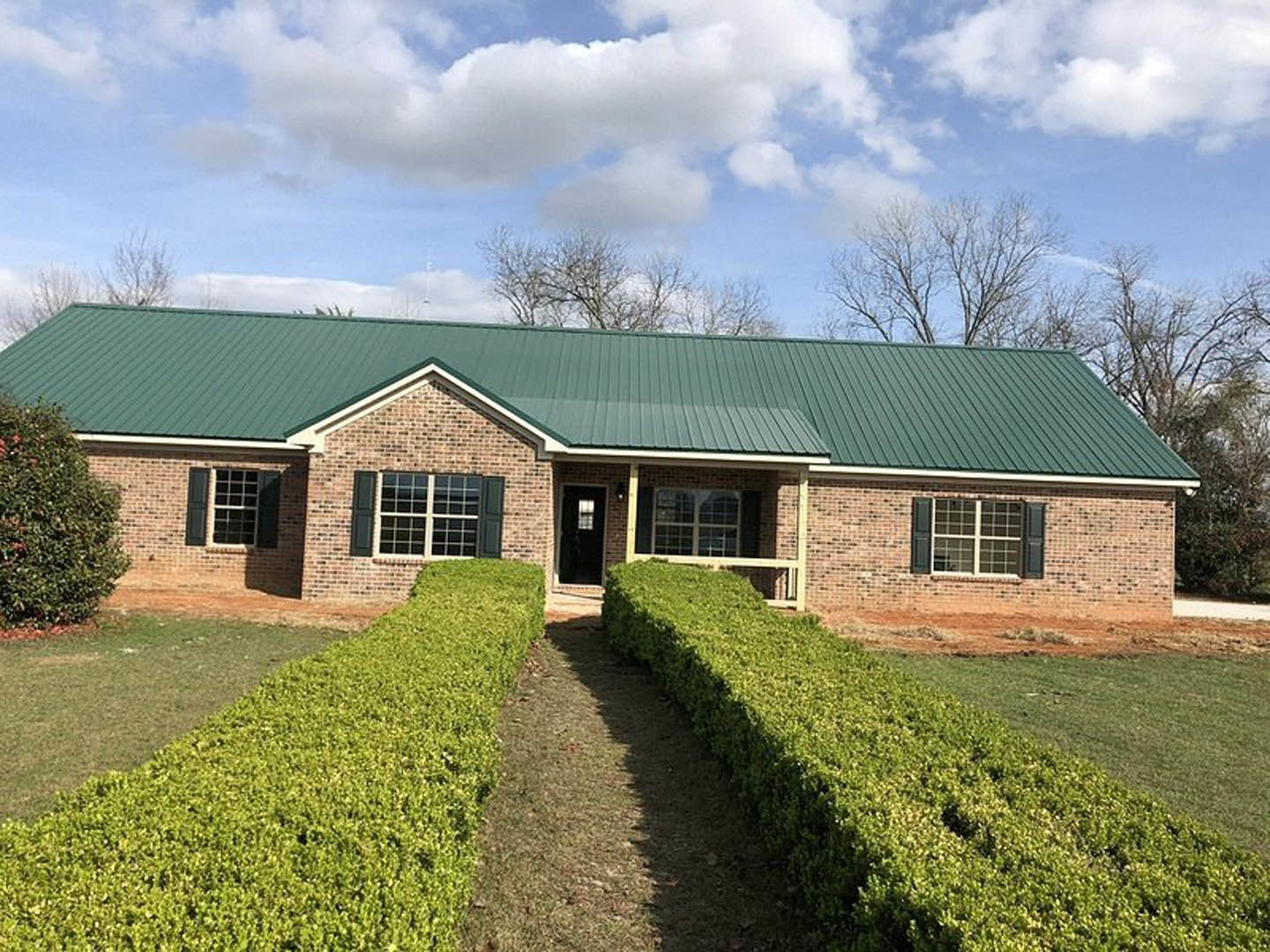 Red brick house with green metal roof, multi-pane windows, trimmed hedge along front, blue sky with scattered clouds