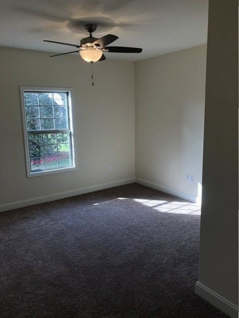 Ceiling fan with light fixture mounted on white plaster ceiling above dark carpeted floor, large window offering garden view, neutral walls