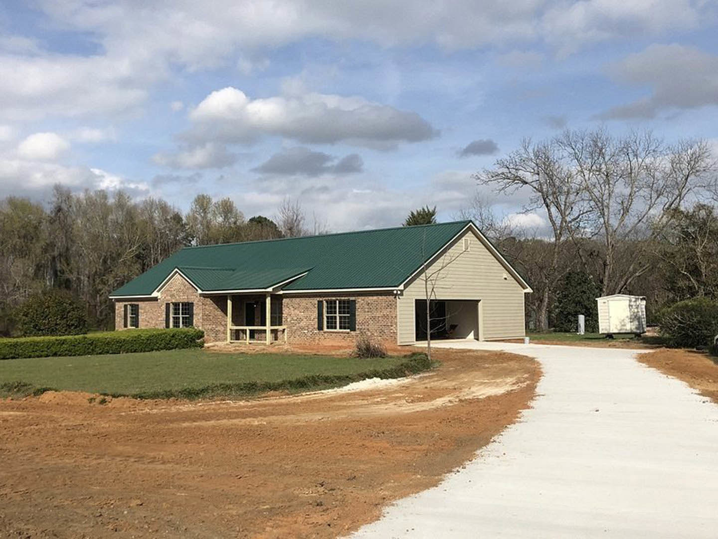 Green metal roof atop a white house with white-framed windows, attached garage, small white shed, concrete driveway, grassy lawn, and partly cloudy sky.