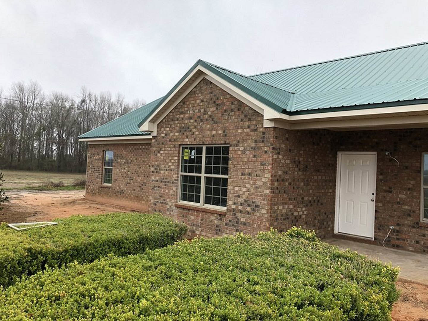 Red brick house with green roof, white front door with silver handle, window displaying yellow sign, white pipe attached to brick wall, bushes lining exterior
