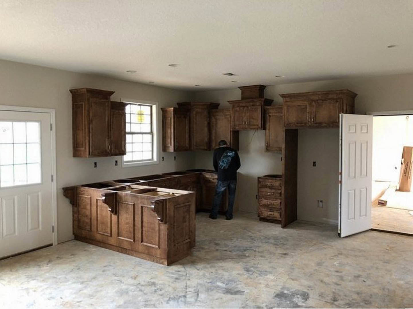Man standing beside wooden kitchen island with drawers, white cabinetry, white door with window, light-colored countertops, and window displaying yellow sign
