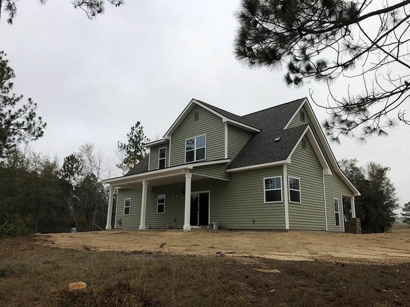Two-story home with wide covered front porch, white trim windows, gray siding, landscaped lawn, and mature trees