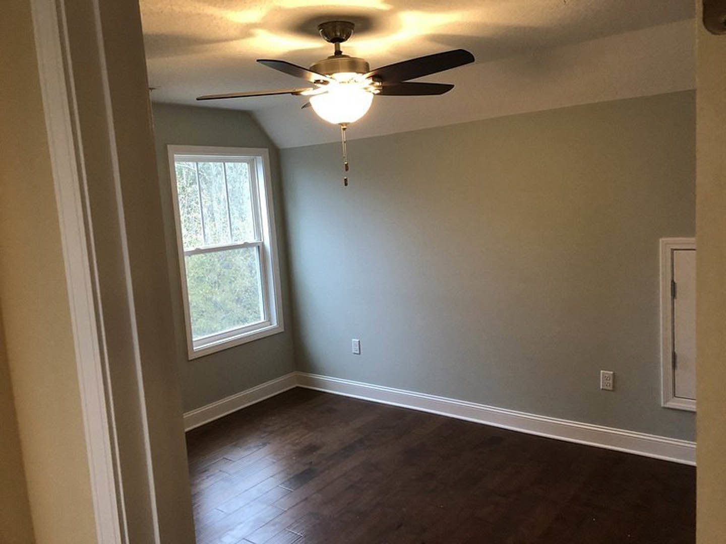 Ceiling fan with light fixture mounted on white plaster ceiling, dark wood floor with white baseboard trim, window with white frame, partial view of white door