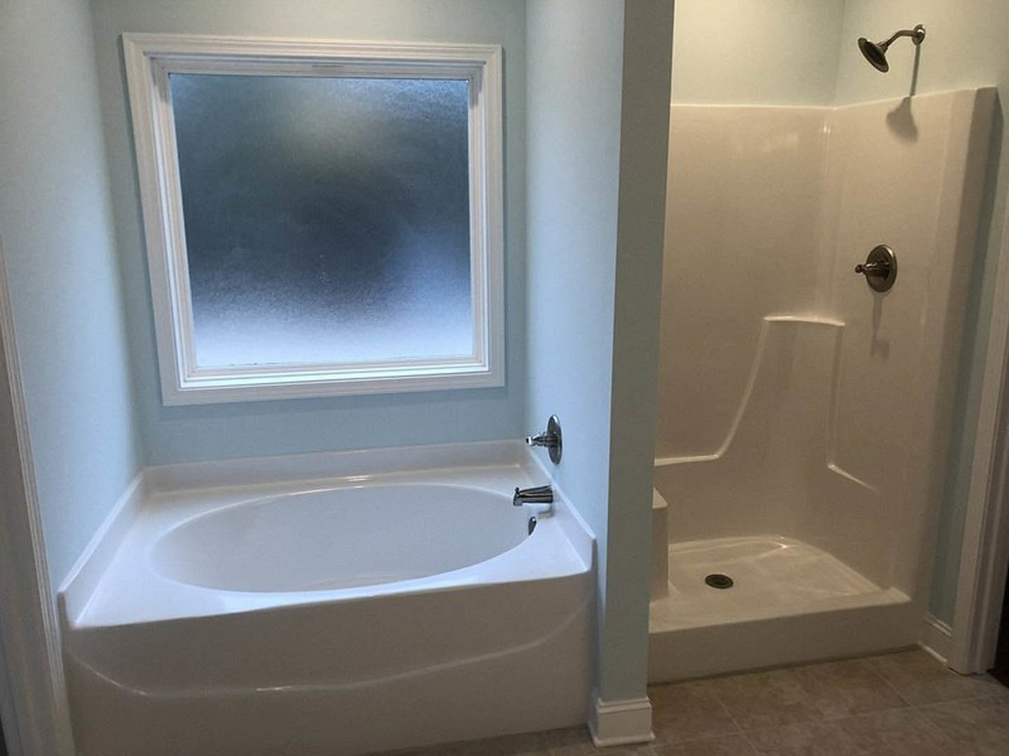 White bathtub with rounded center next to glass-enclosed shower with silver handle, frosted window above, light tile walls, chrome hook, and bathroom accessories visible.