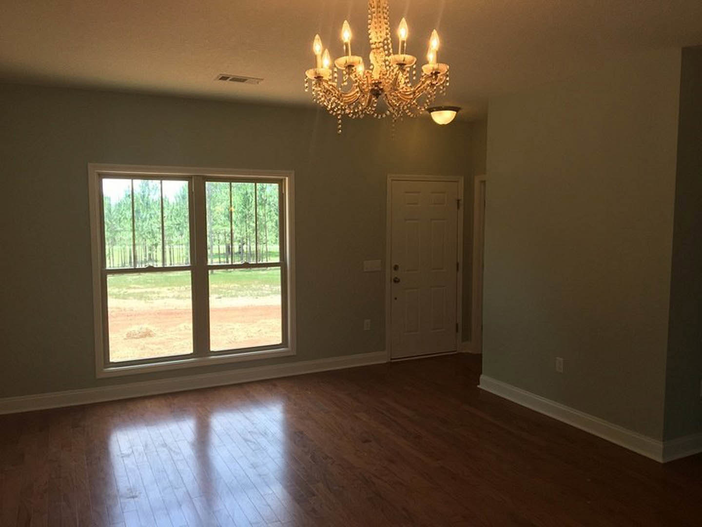 Hardwood floor room with multi-light chandelier, white paneled door, large window showing trees, crown molding, and soft natural light