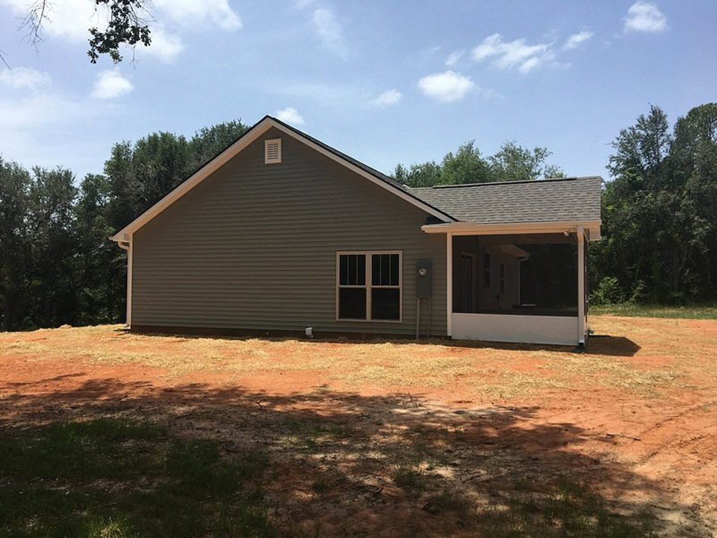 Two-story house with white-framed windows, covered front porch, gray siding, shingled roof, grassy yard, and mature trees in the background