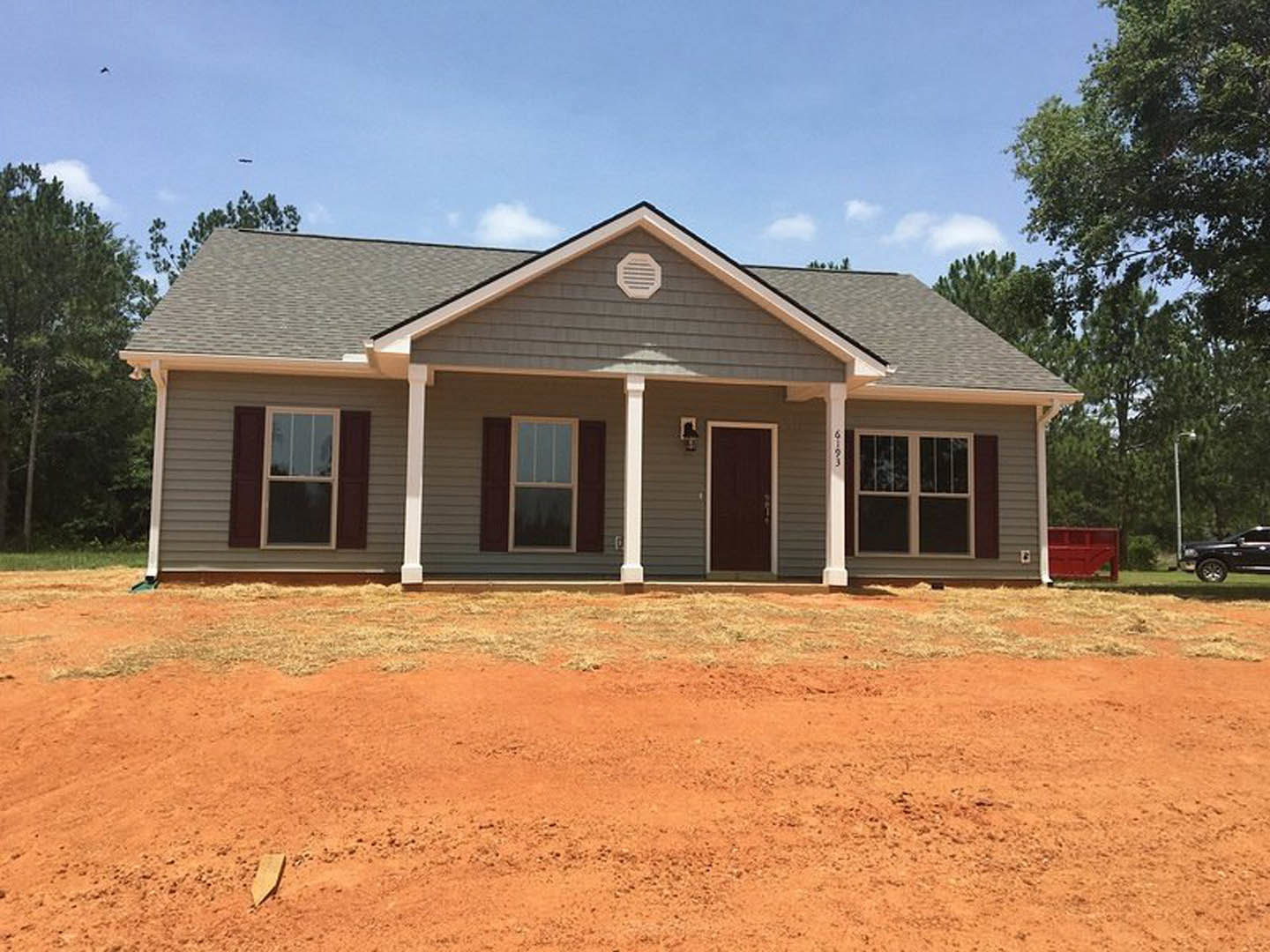 Single-story house with gray roof, white siding, dark brown door with white trim, white wall vent, and large front window; dirt area in front yard, black truck parked on grass