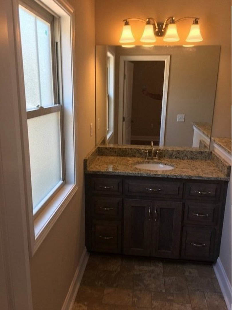 Bathroom with brown tile floor, white sink set in a wood cabinet, rectangular mirror above, two-light wall fixture, partial view of a hand in the doorway.