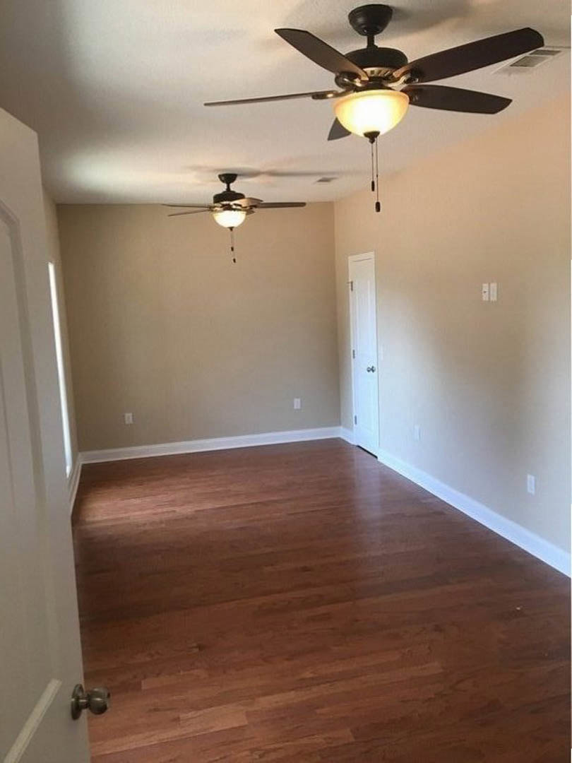 Spacious room with warm wood flooring, white walls, ceiling fan with light fixture, white door with metal handle, floor vent near baseboard