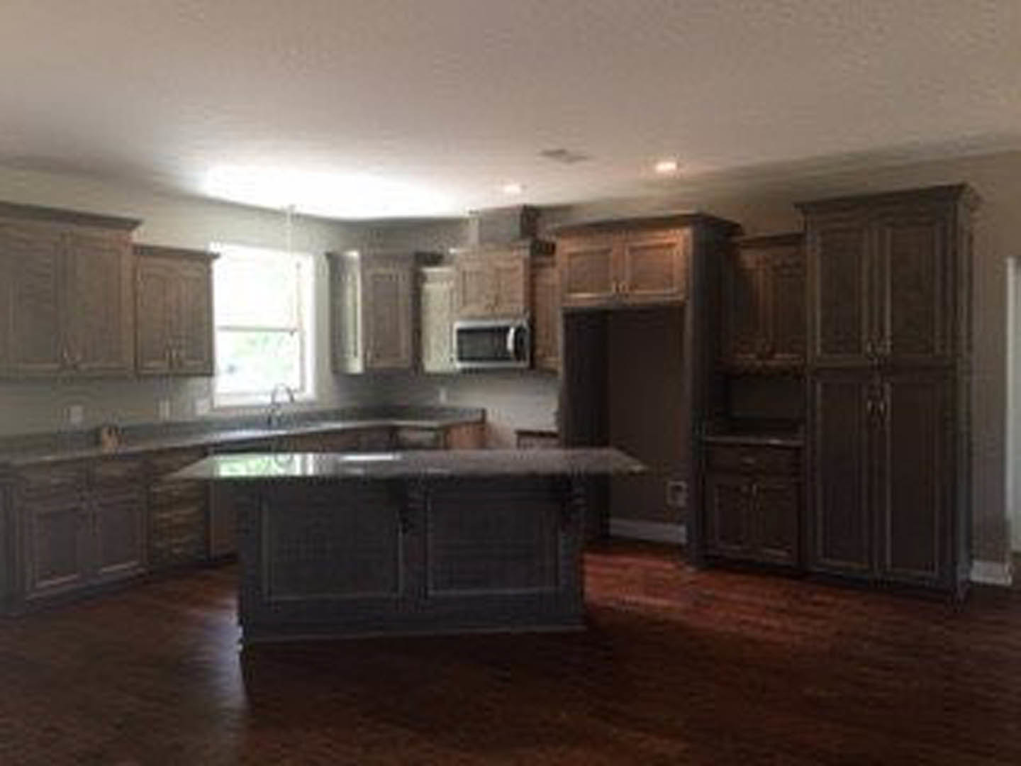 Spacious kitchen featuring a large granite island, black cabinetry, light wood flooring, and stainless steel appliances; a person walks past in the background.