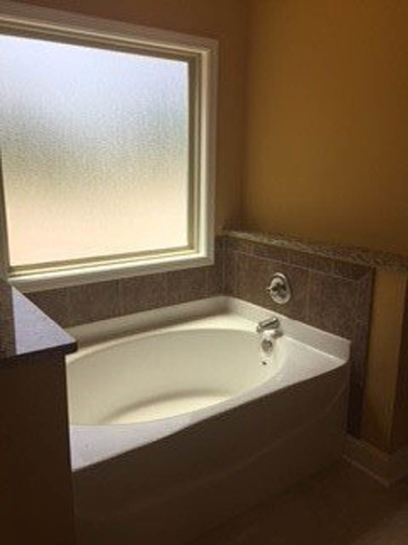 White freestanding bathtub with silver fixtures beside frosted glass window, white sink and brown countertop, light tile walls and floor