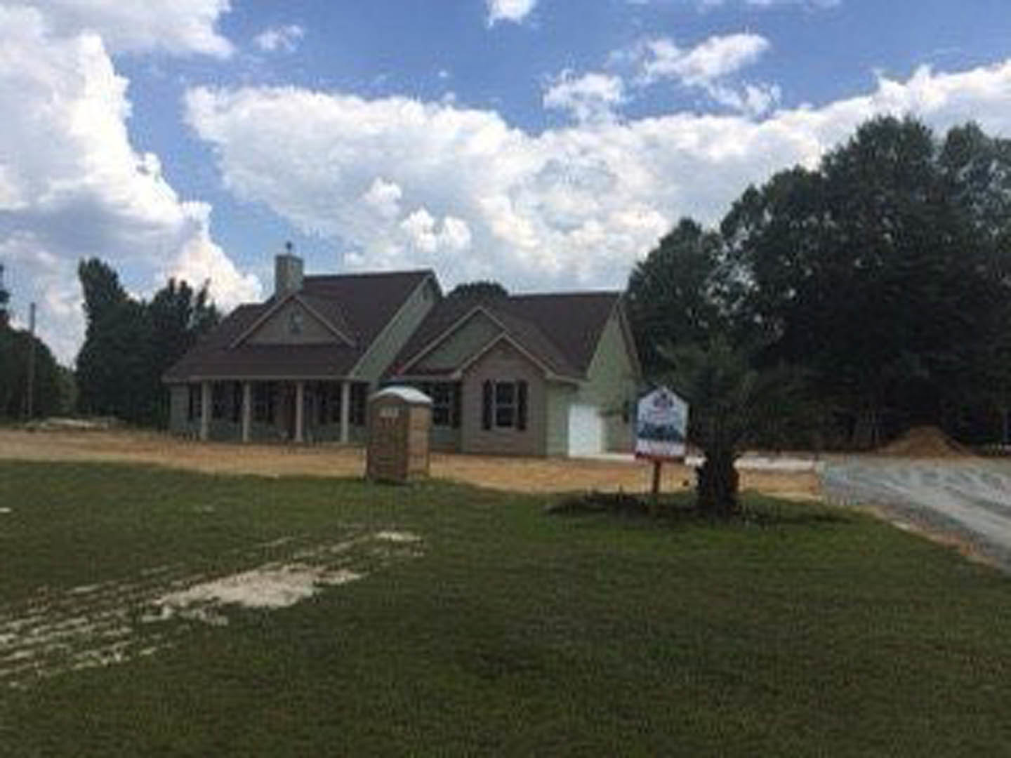 Two-story custom home with white siding, black-framed windows, gabled roof, manicured lawn, and a real estate sign near the walkway; trees and clouds in the background.