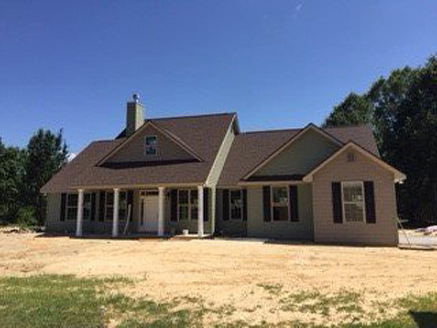Two-story house with gray siding and white trim, covered front porch, dirt yard in foreground, mature trees surrounding property, cloudy sky overhead