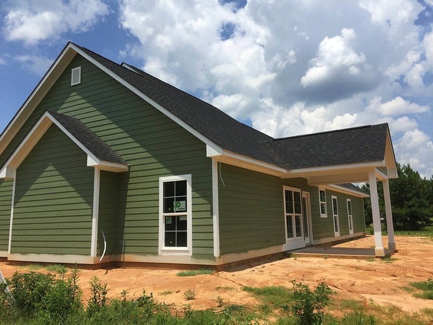 Green siding with white trim, multiple windows including one with a sign, dirt patch in front yard, leafy tree to the side, gray shingled roof under partly cloudy sky