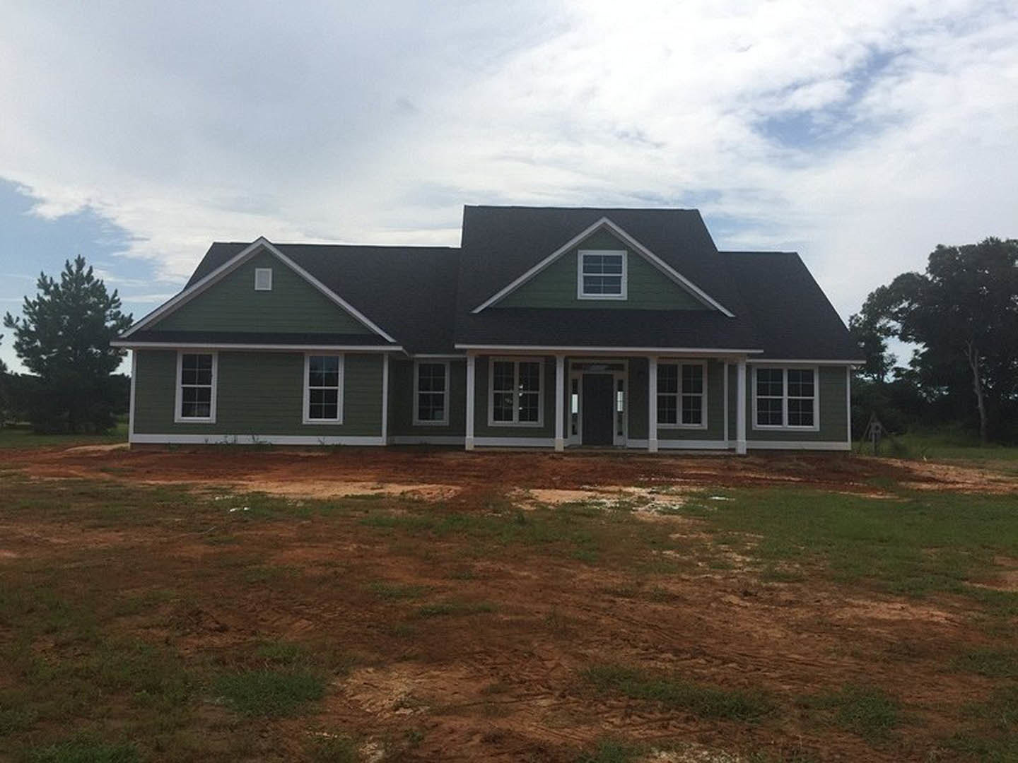 Two-story custom home under construction with black roof, white-framed windows, and covered porch; surrounded by dirt, grass, and leafy trees under a partly cloudy sky