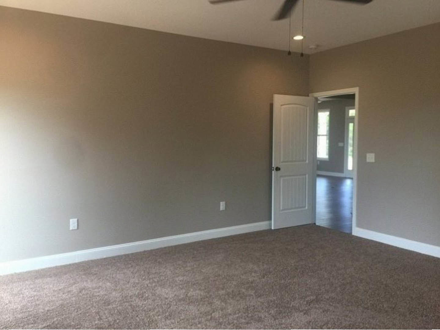 White paneled door open to a carpeted room with white baseboards, ceiling light fixture, and blurred hand near the doorway