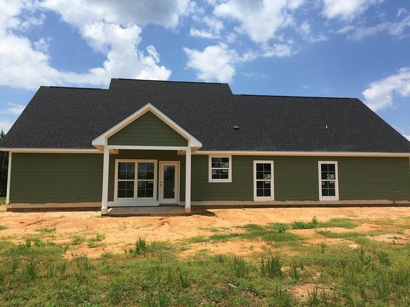 Two-story house with green metal roof, green siding, white trim, multi-pane windows, white glass front door, grassy and dirt yard in foreground, cloudy sky above