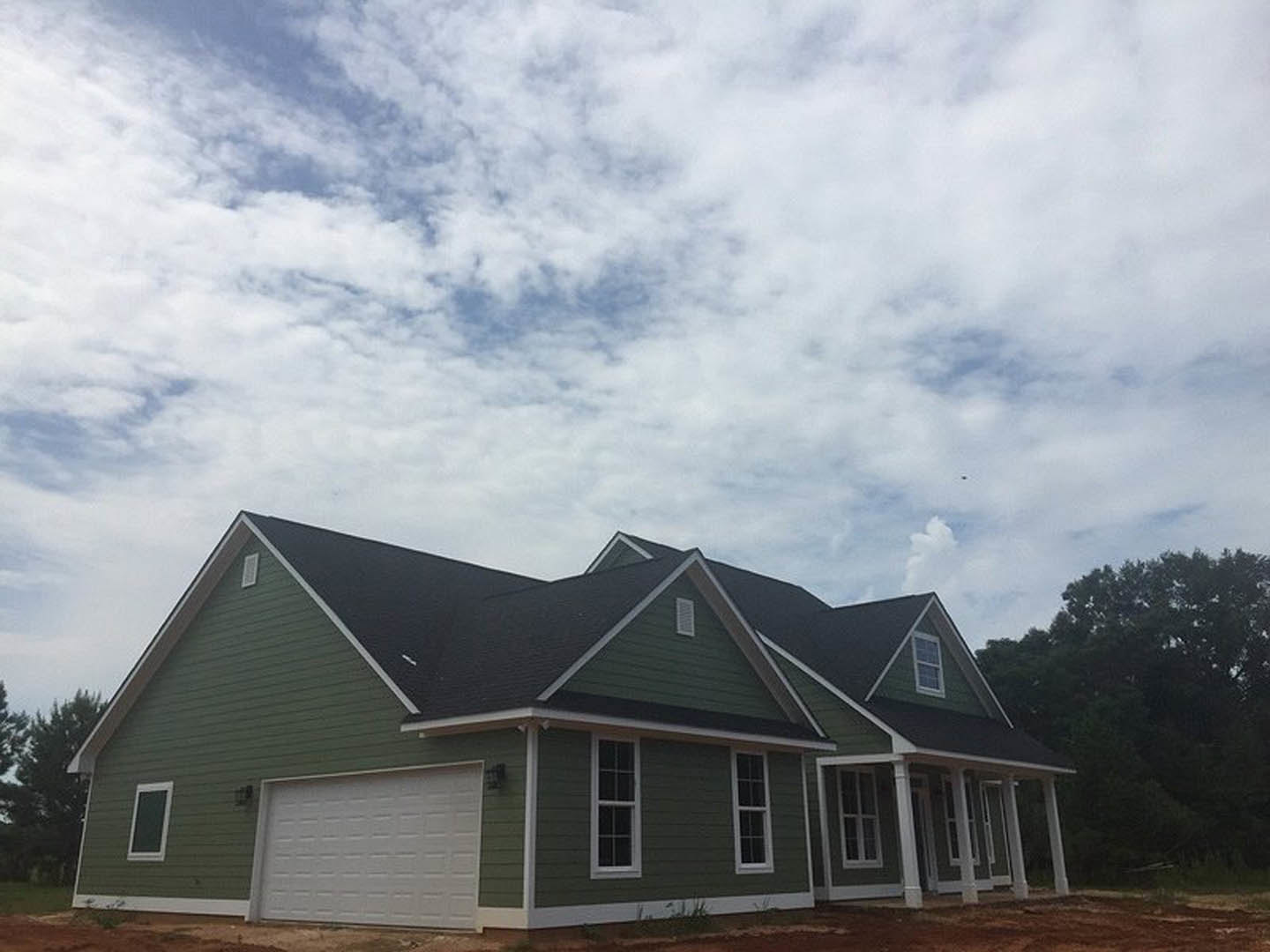 White garage door with green trim, light-colored siding, gabled roof, mature trees in background, cloudy sky overhead