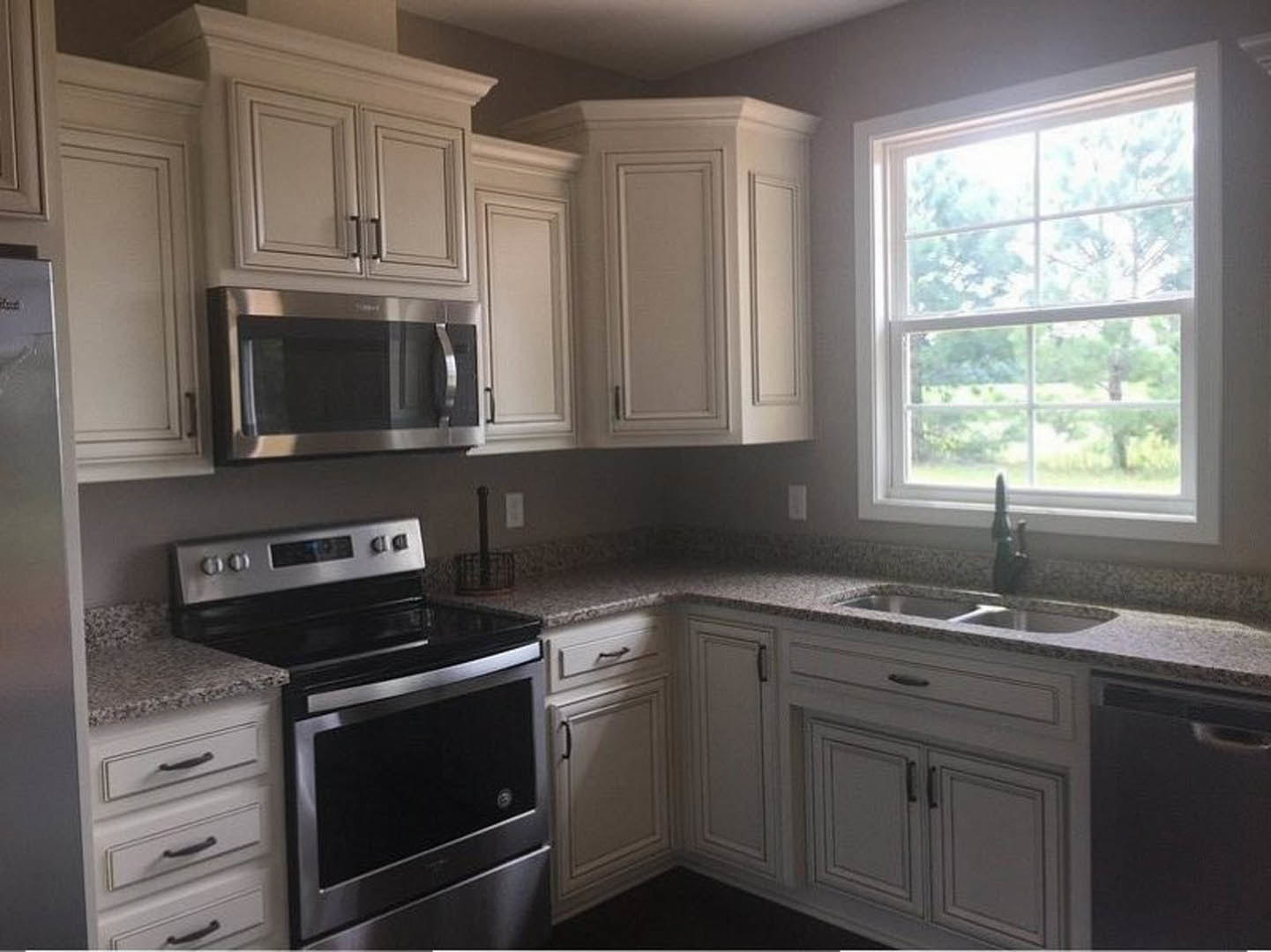 White kitchen cabinets with granite countertops, stainless steel stove and microwave, framed black and white artwork on white wall, window overlooking green trees