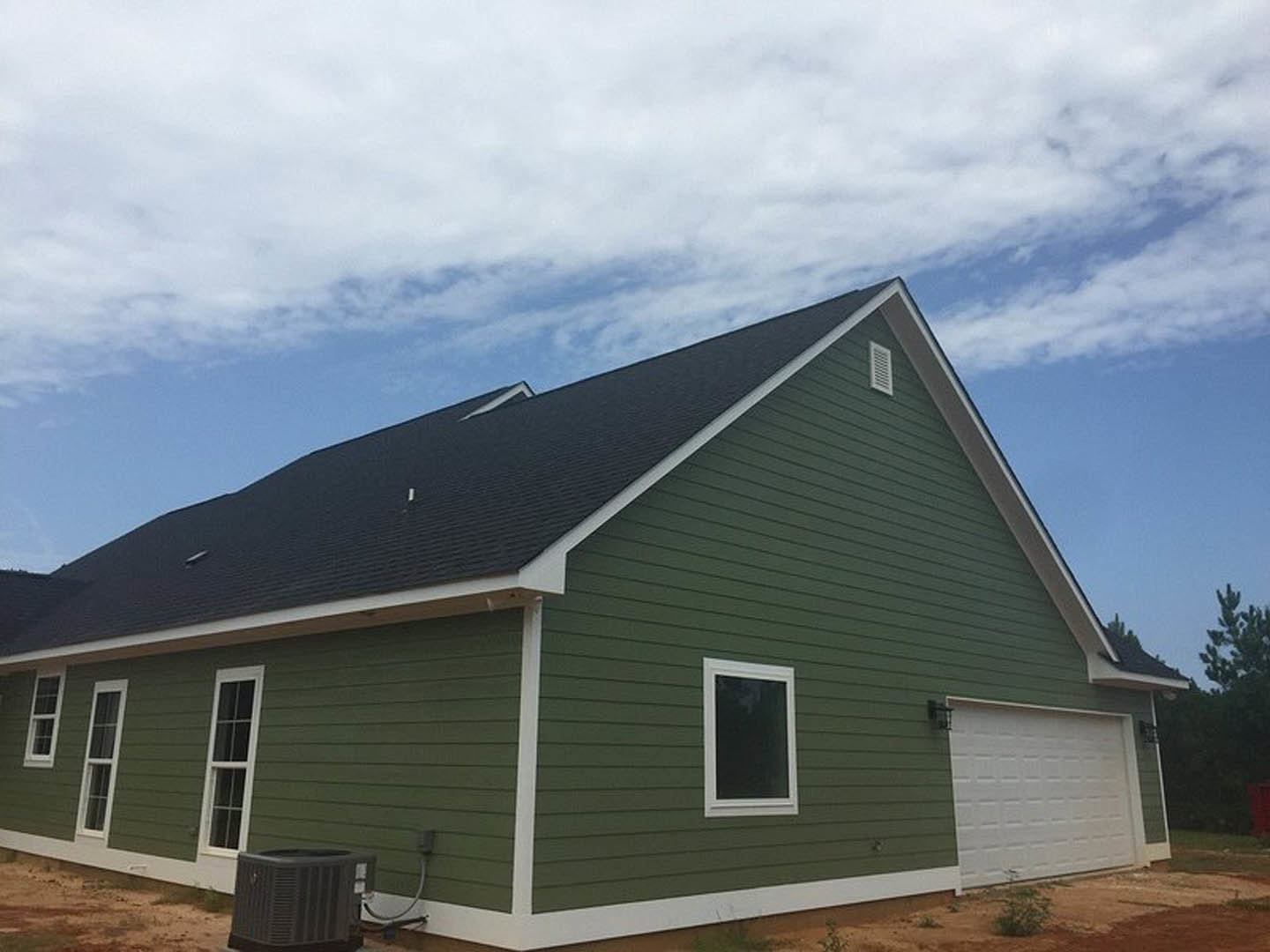 Green house with black roof, white-framed windows, attached garage with paneled door, surrounded by grassy yard under cloudy sky