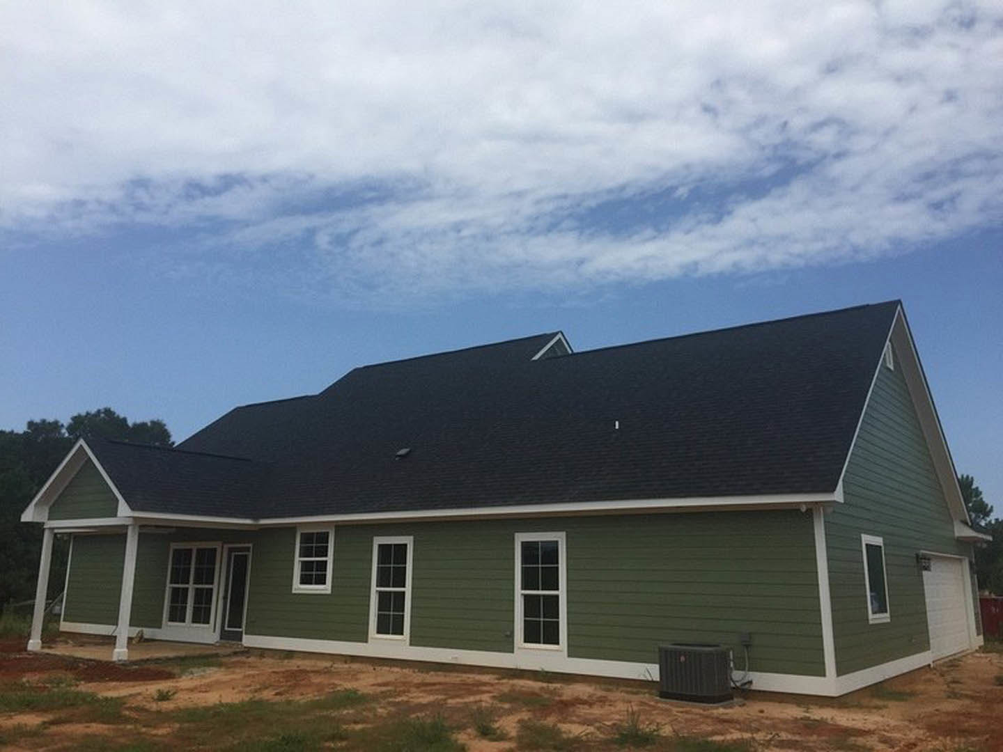 Green siding house with white trim, white-framed windows, black rectangular mailbox with red logo, blue sky with scattered clouds, dirt patch in front yard, close-up of shingled
