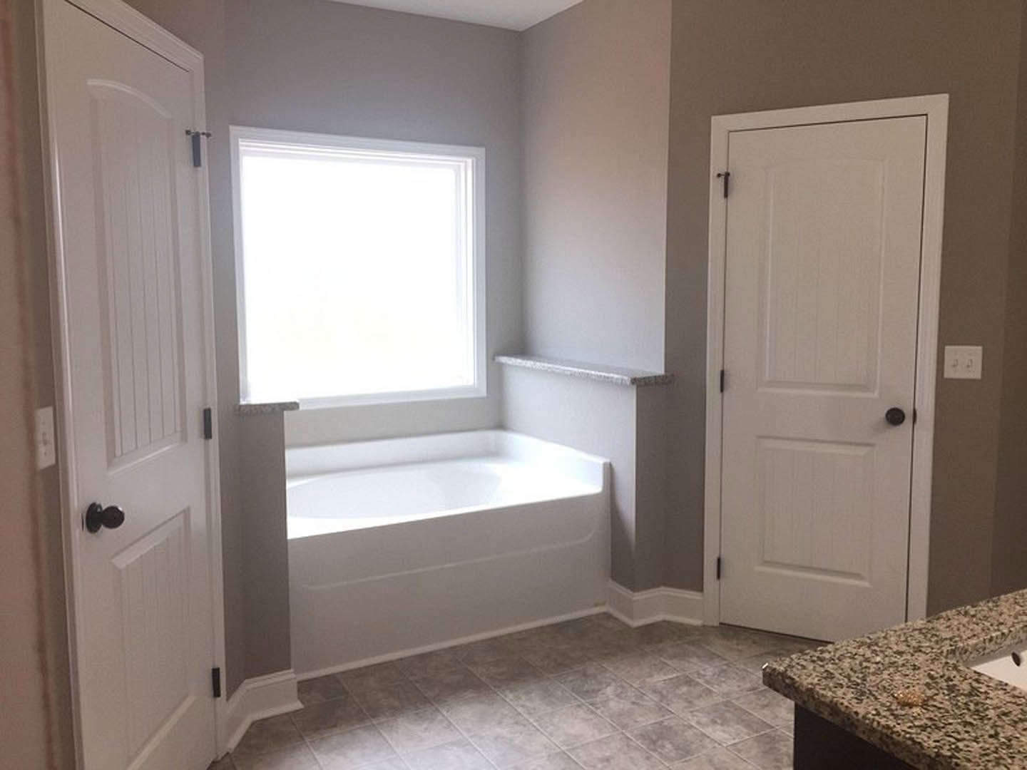 Bathroom with freestanding bathtub, marble countertop and sink, two doors, white walls, grey tile flooring, rectangular window, wall switch, and black decor object on counter