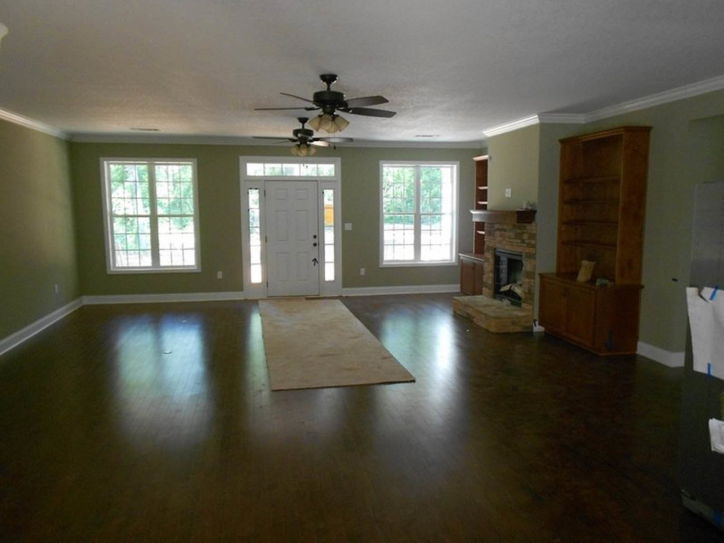 Living room with laminate wood flooring, white plaster walls, ceiling fan with light, central fireplace, wooden shelf holding a bag, brown mat on floor, white door with black