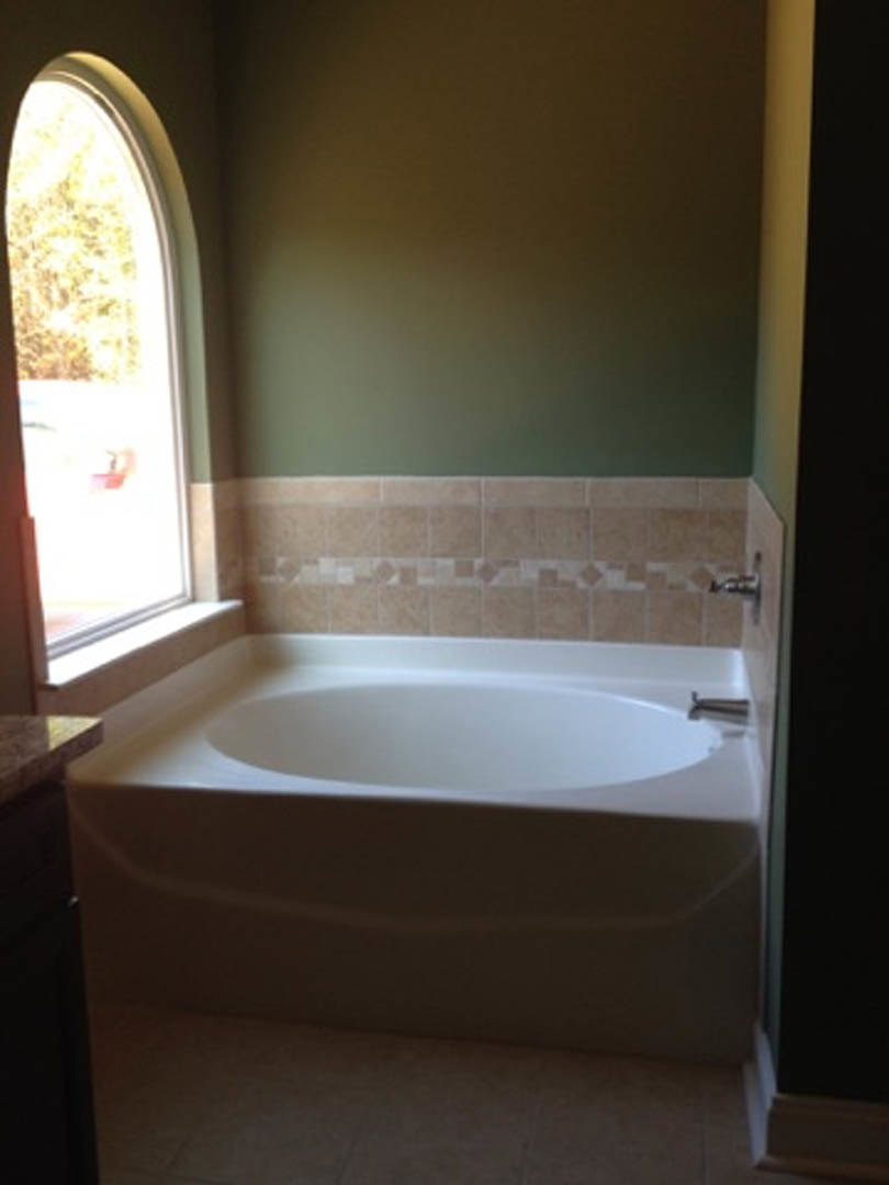 Freestanding white bathtub with chrome faucet beside large window, surrounded by light gray tile walls and floor