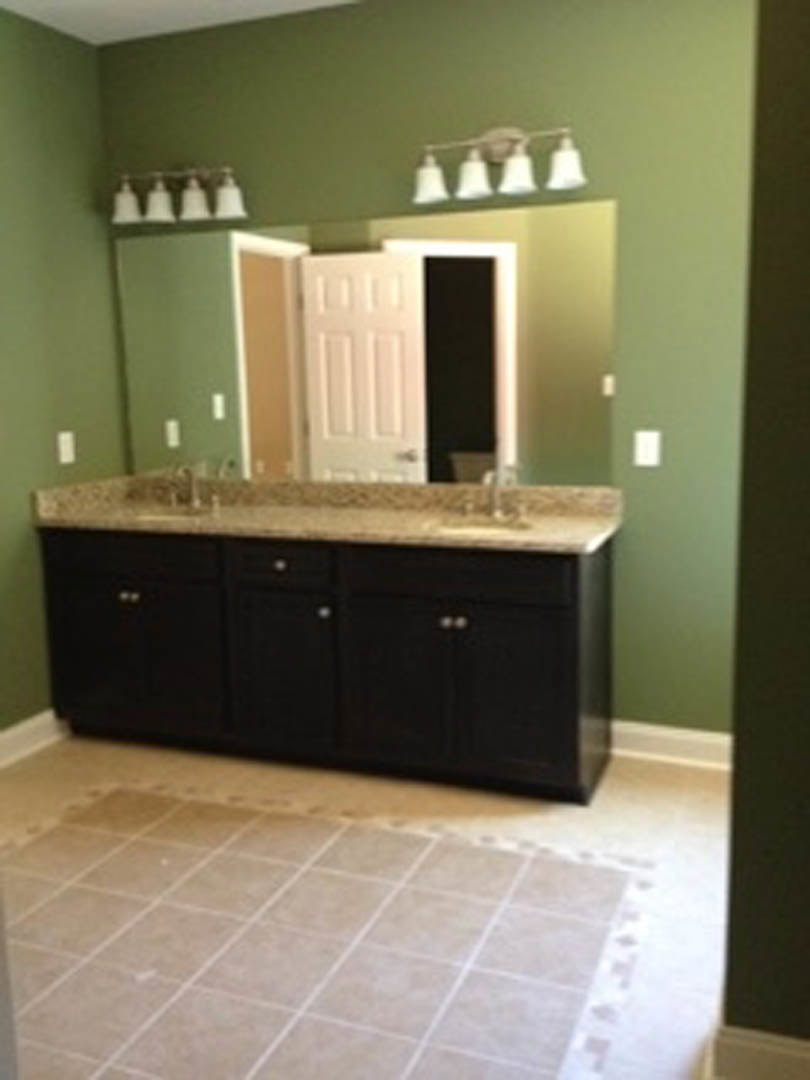 Double vanity with white sinks and chrome faucets, quartz countertop, green accent wall, tile floor, white cabinetry, and open door in a modern bathroom.