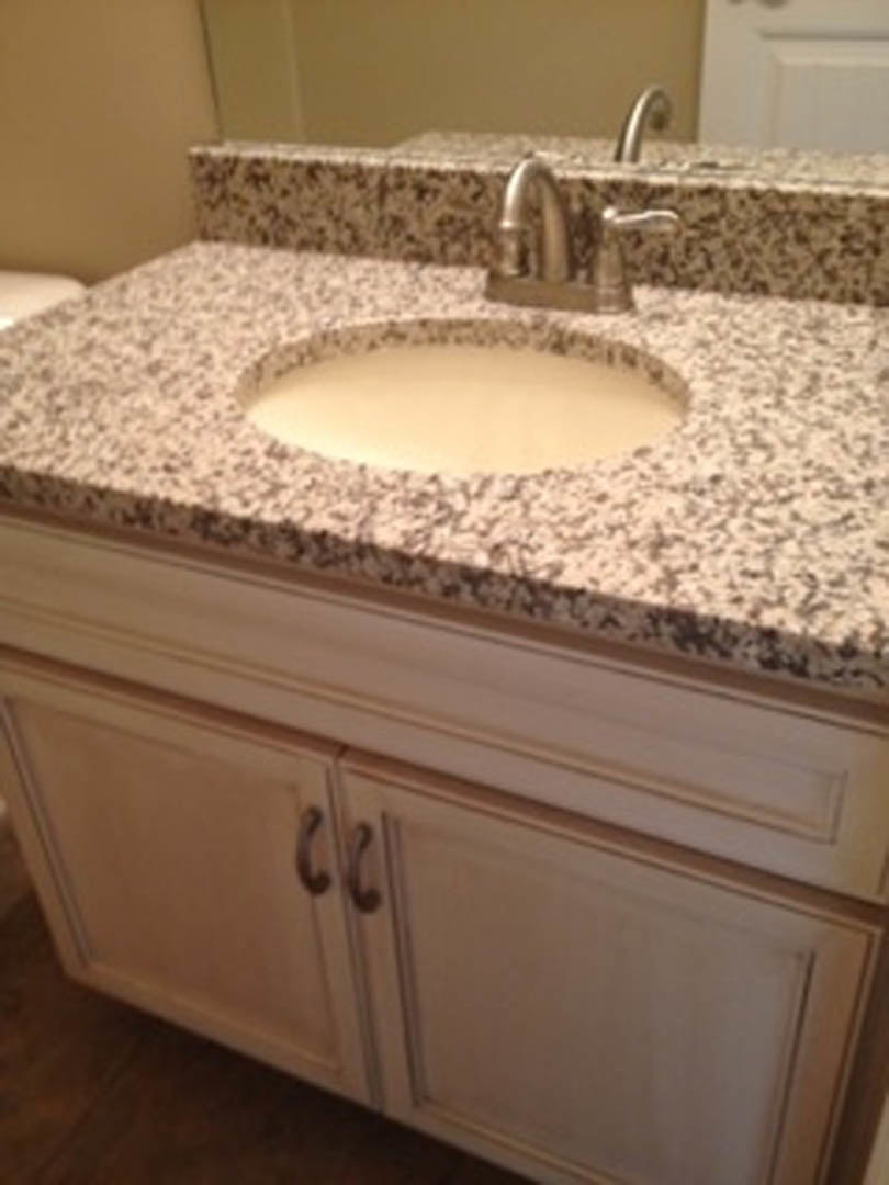 White bathroom sink with chrome faucet set in a light countertop, adjacent to white cabinetry and a tiled wall.
