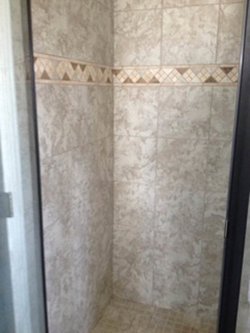 Walk-in shower with clear glass door, marble tile walls, and tile floor; partial view of window and person's hand in foreground