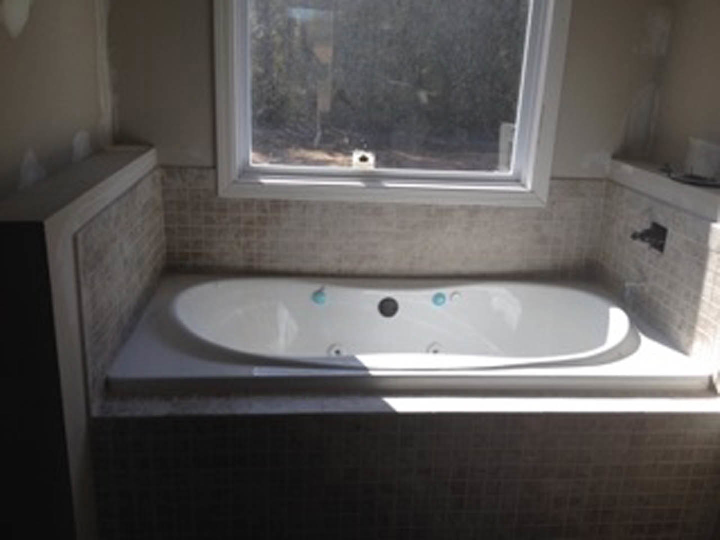 Freestanding white bathtub with chrome faucet, surrounded by light tile flooring and wall, large window and mirror reflecting natural light