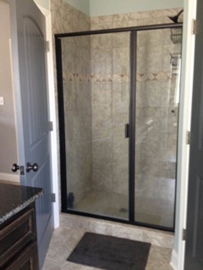 Walk-in shower with frameless glass doors, white tile walls, black vanity countertop, chrome fixtures, and built-in cabinetry