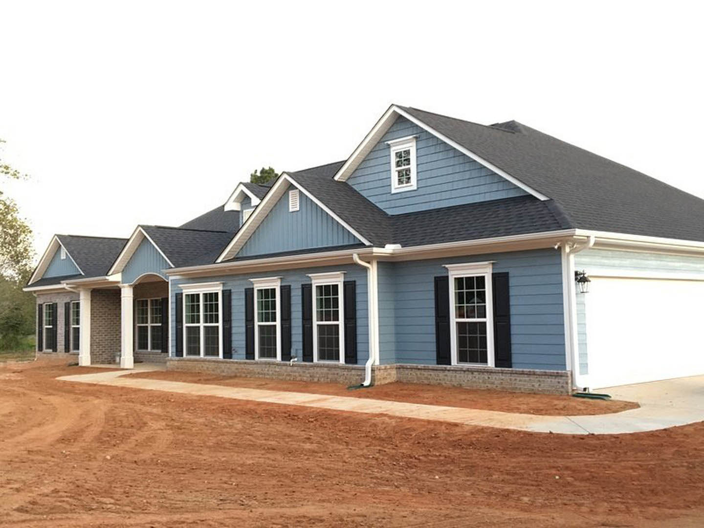 Blue house with black shutters and white-framed windows, dirt road in foreground with white line, tree visible to the side, cloudy sky above