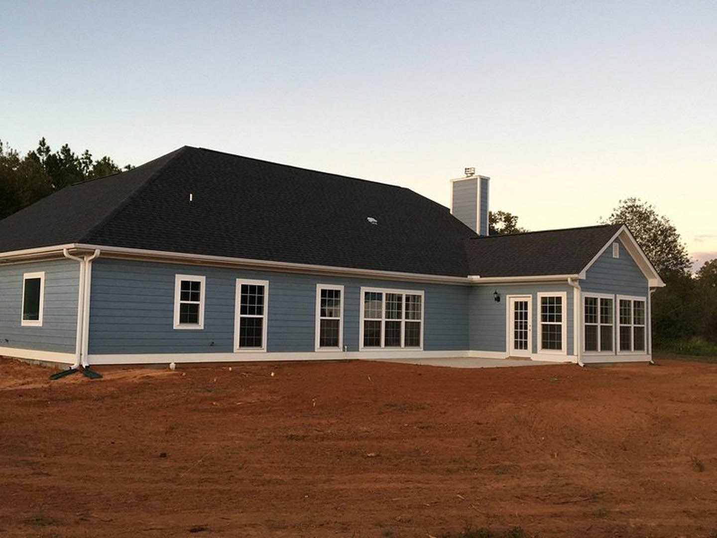 Blue siding house with white trim and large windows, set behind a spacious brown dirt yard, under clear sky.