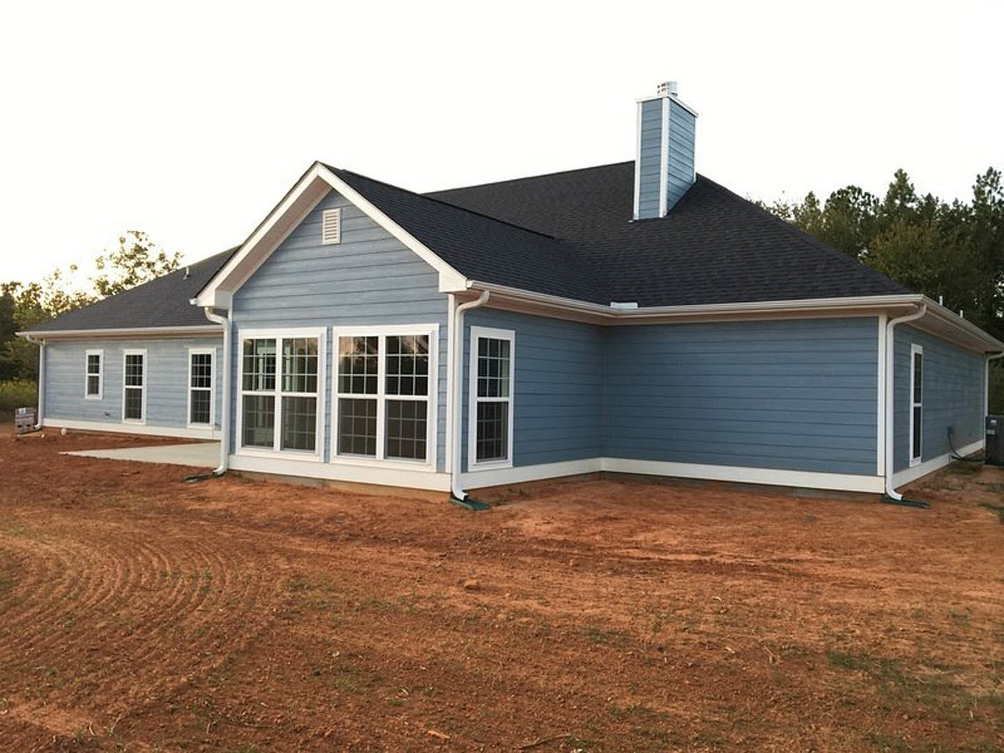 Tan siding house with white-framed windows, brick chimney, and bare dirt yard under a clear sky