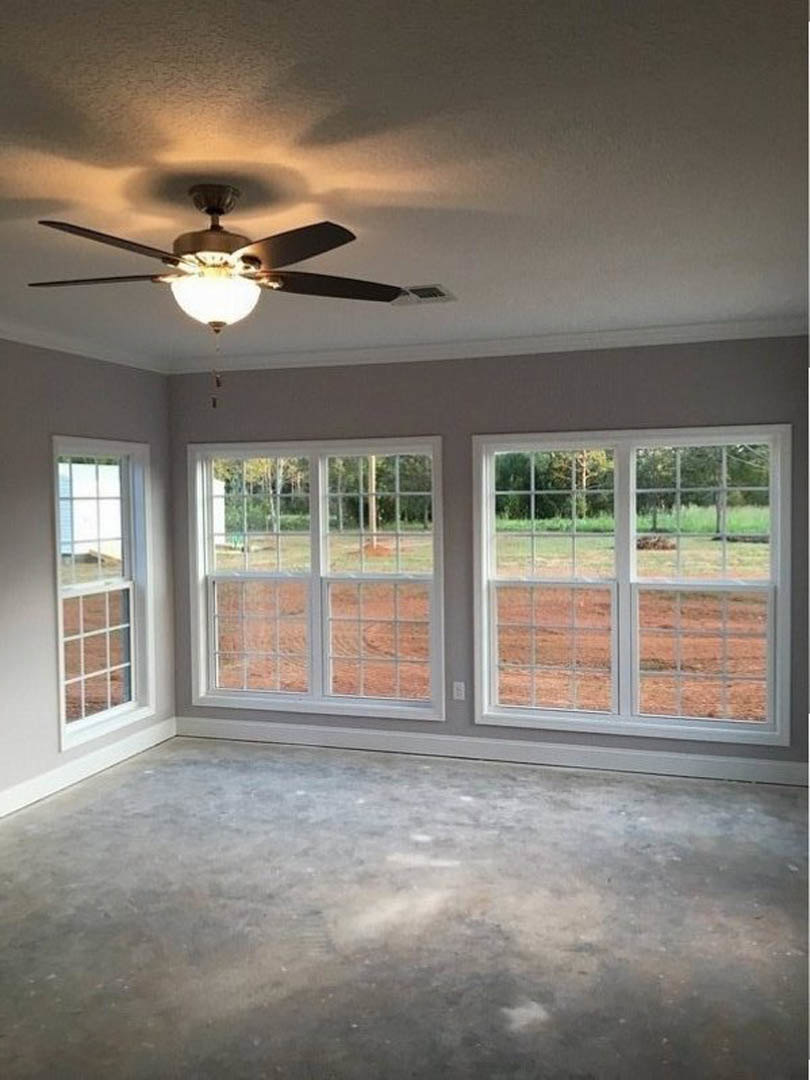 Ceiling fan with light fixture mounted on white ceiling, large windows with views of grassy field, smooth concrete floor, neutral walls, natural daylight illuminating modern