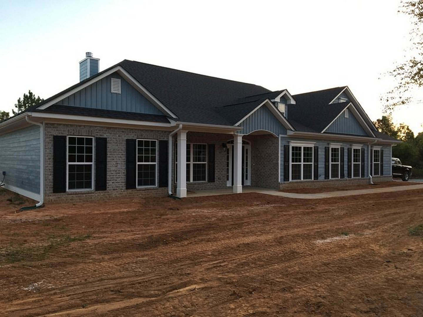 Brick and stone house with white columns, grid windows, and a black truck parked on a dirt road in front, surrounded by trees and sky.