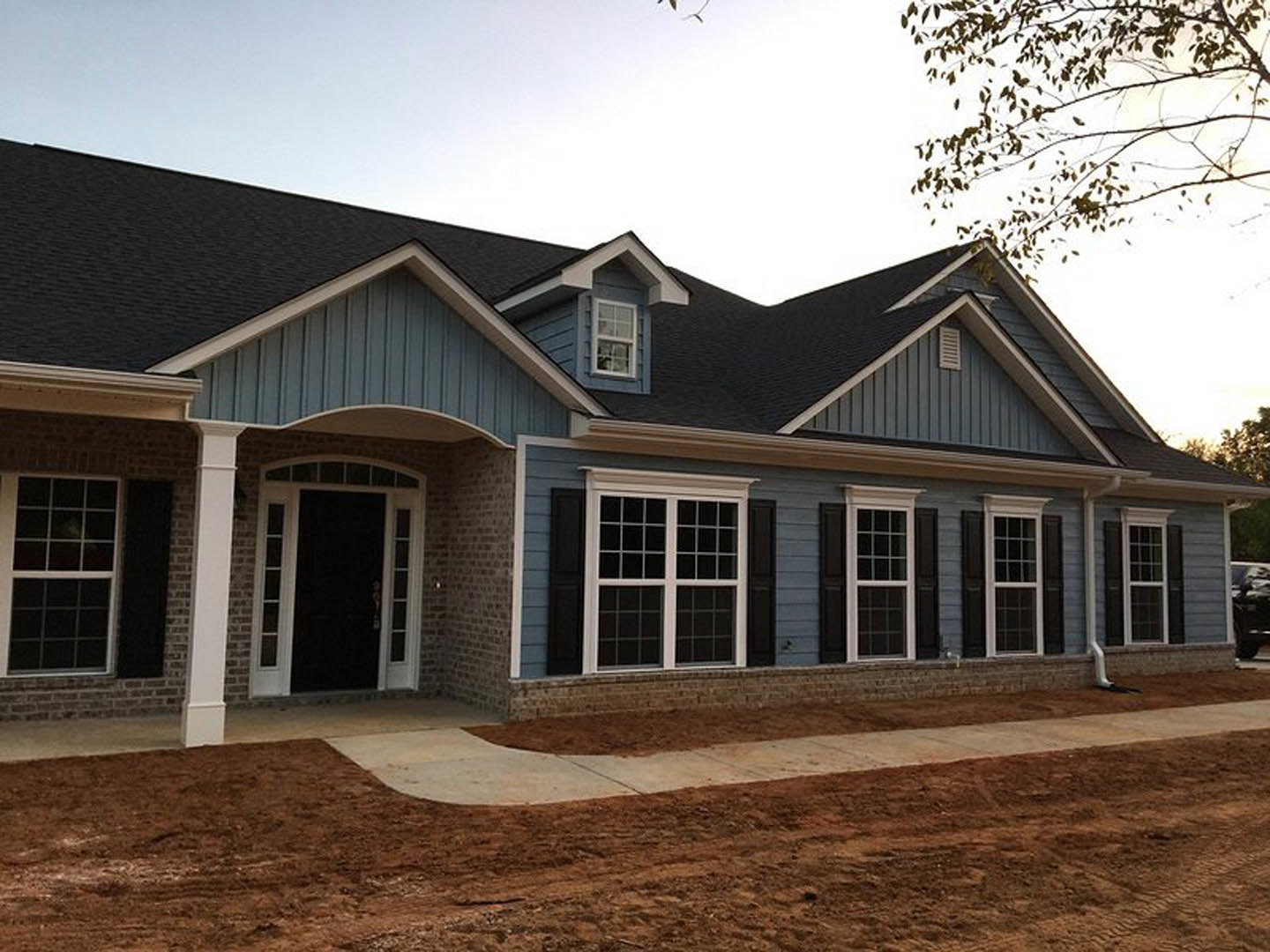 Black front door with white trim, white-trimmed window, light-colored siding, dirt driveway and path, leafy tree branches in foreground