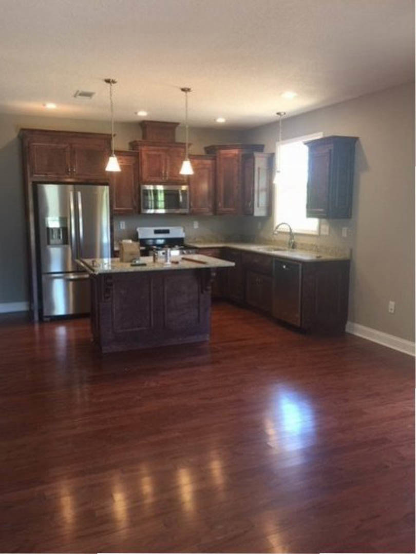 Kitchen with shiny wood flooring, marble-topped island, stainless steel refrigerator, white cabinetry, and blurred window in background.