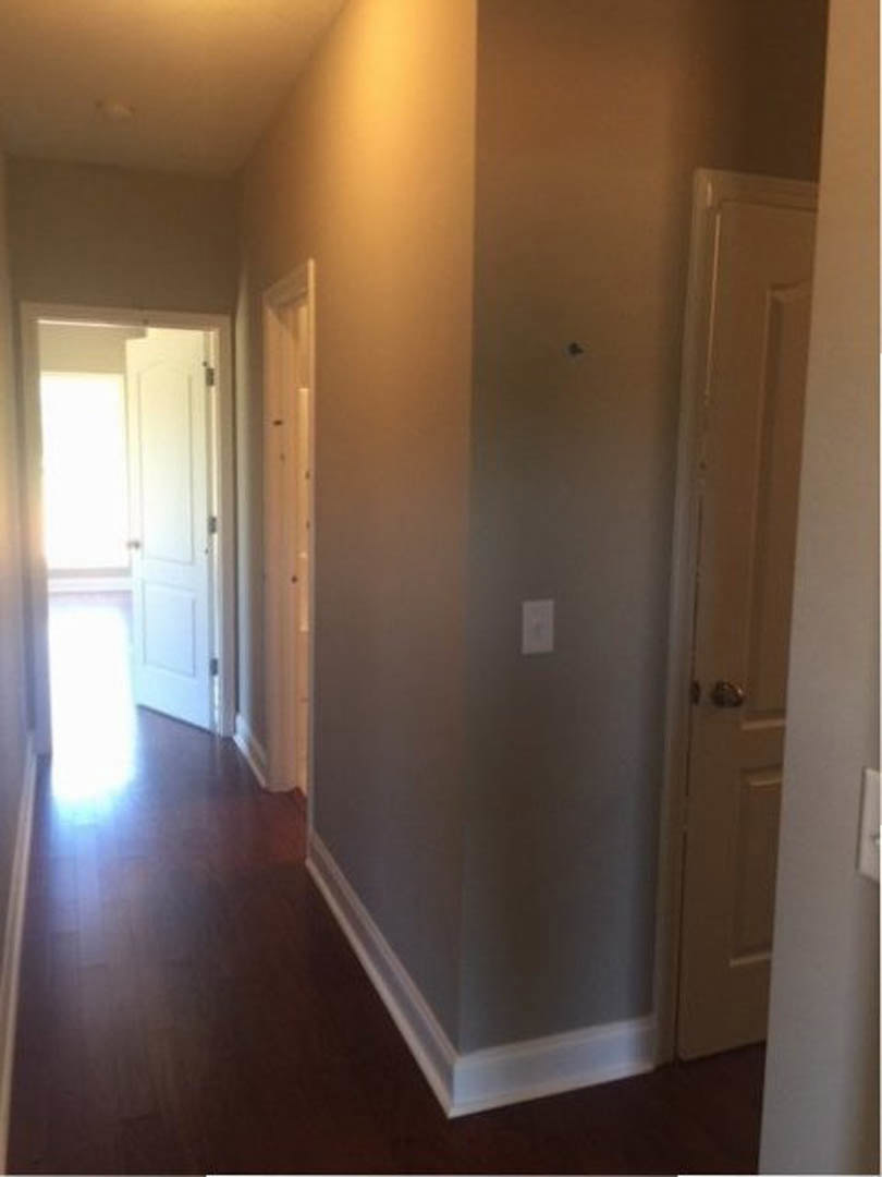 Hallway with light fixture mounted on plaster wall, white outlet and light switch, white door featuring silver door knob, wood flooring