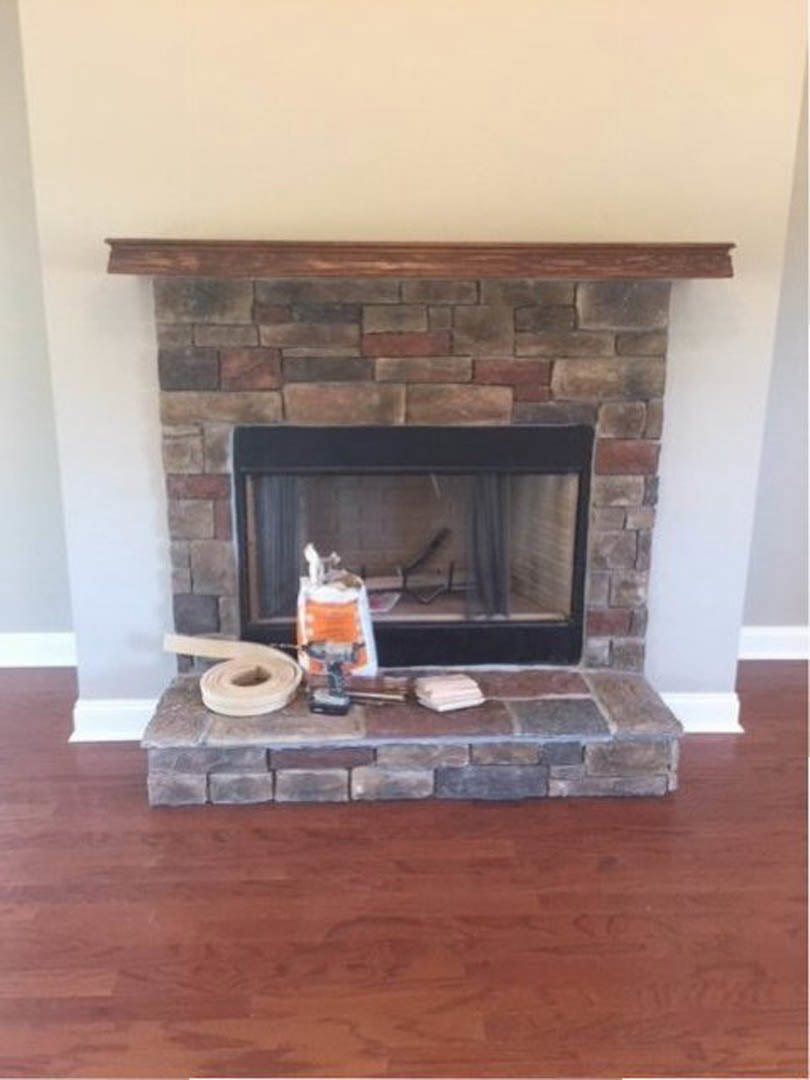 Brick fireplace with black-framed glass window, fireplace tools resting on hearth, wood flooring, and stone wall in background.
