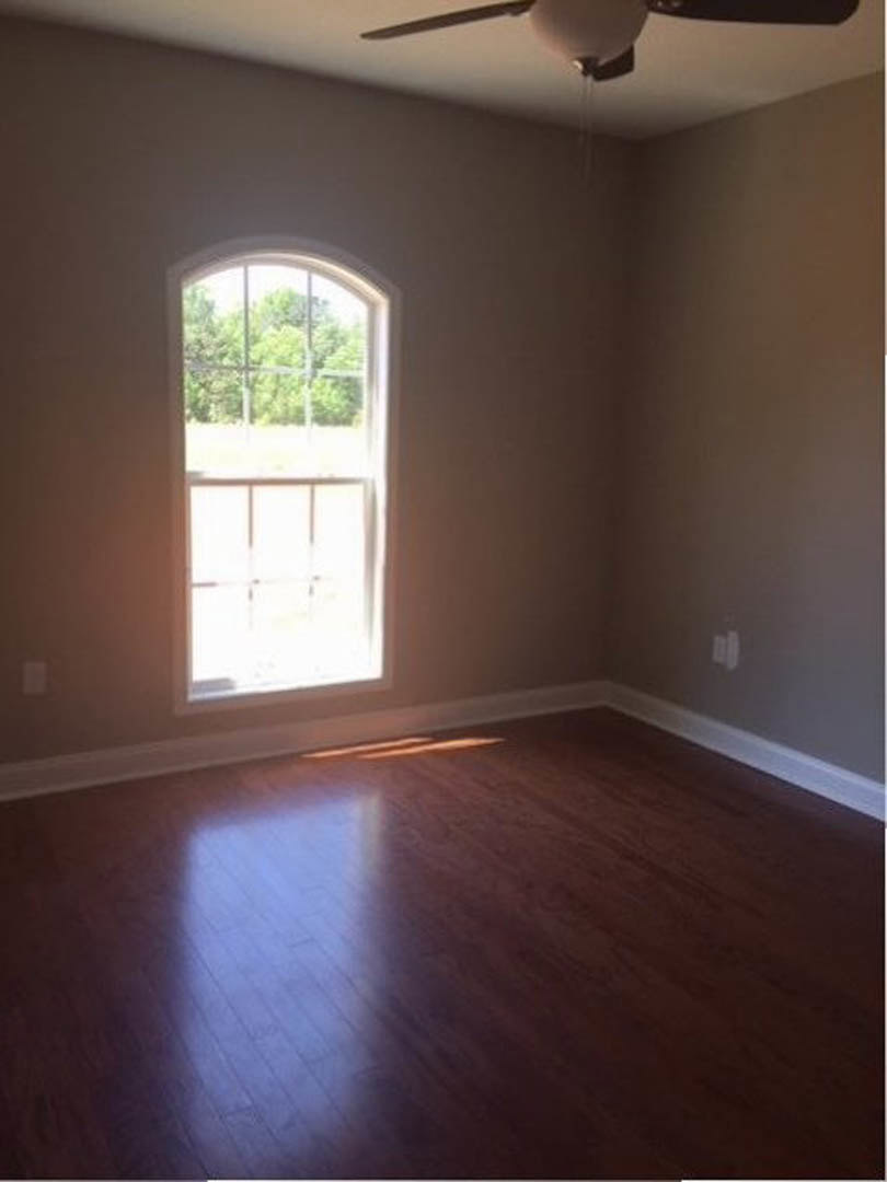 Sunlit room featuring wide-plank hardwood flooring, large window with outdoor view, neutral painted walls, and modern ceiling fan.