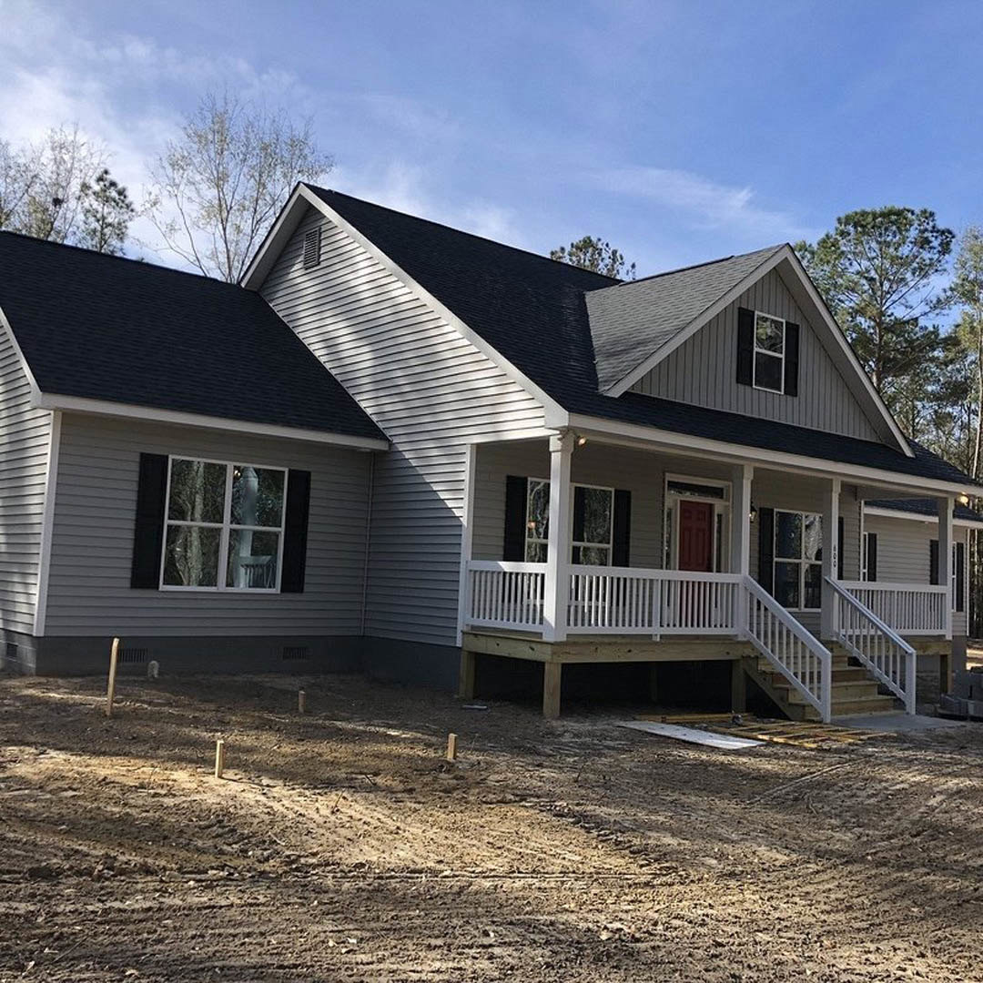 Two-story house under construction with white siding, red front door, white porch railing, white-framed windows, dirt yard, and surrounding trees