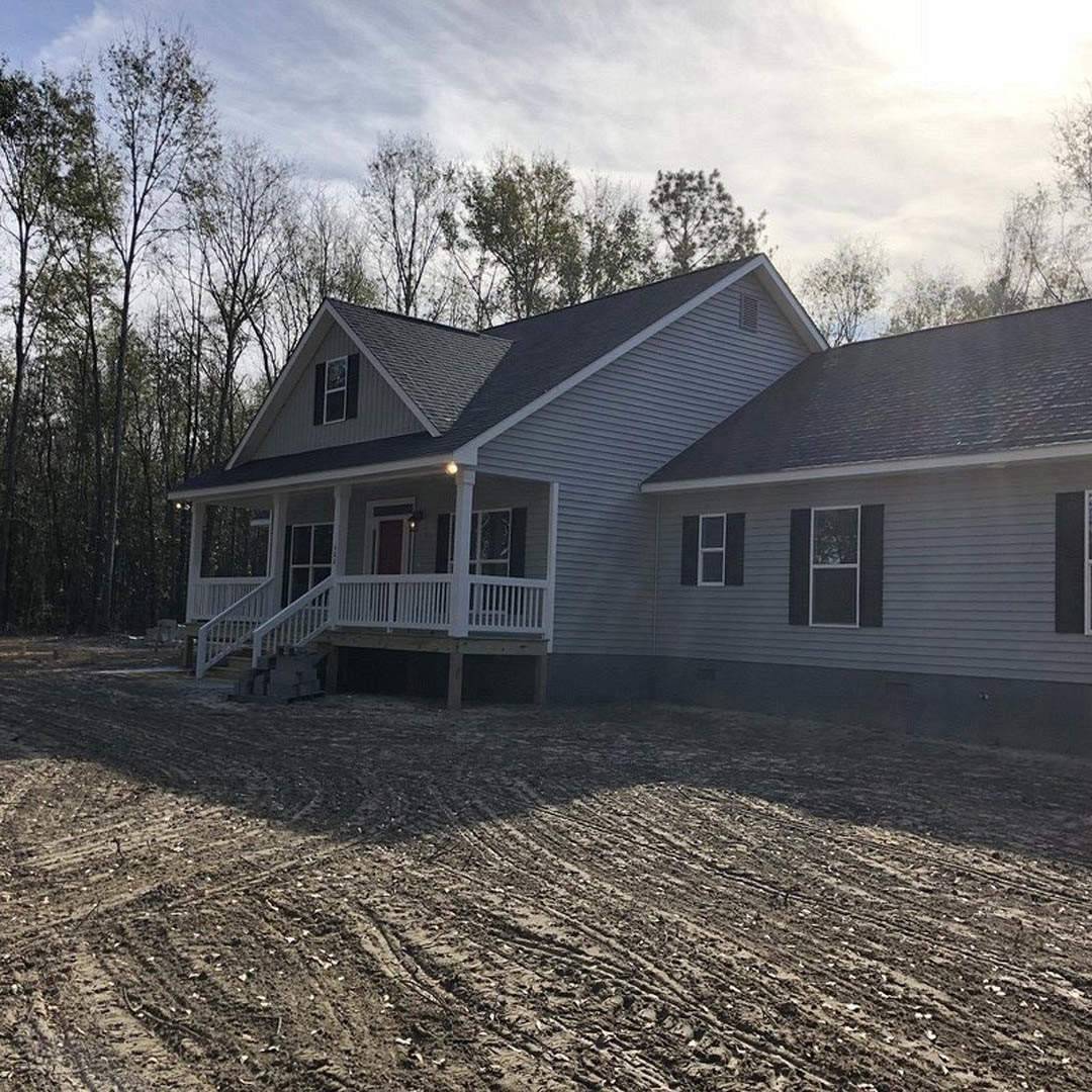 Two-story house with gray siding, white-framed windows, covered porch, white deck railing, and dirt driveway surrounded by trees under a cloudy sky