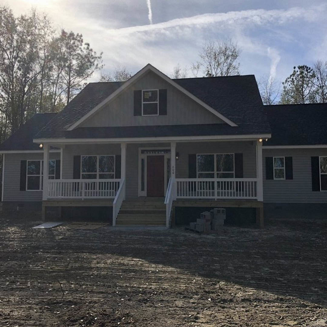 Two-story house with gray siding, covered front porch, white trim, and stairs leading to a dirt yard; large windows and a cloudy sky overhead.