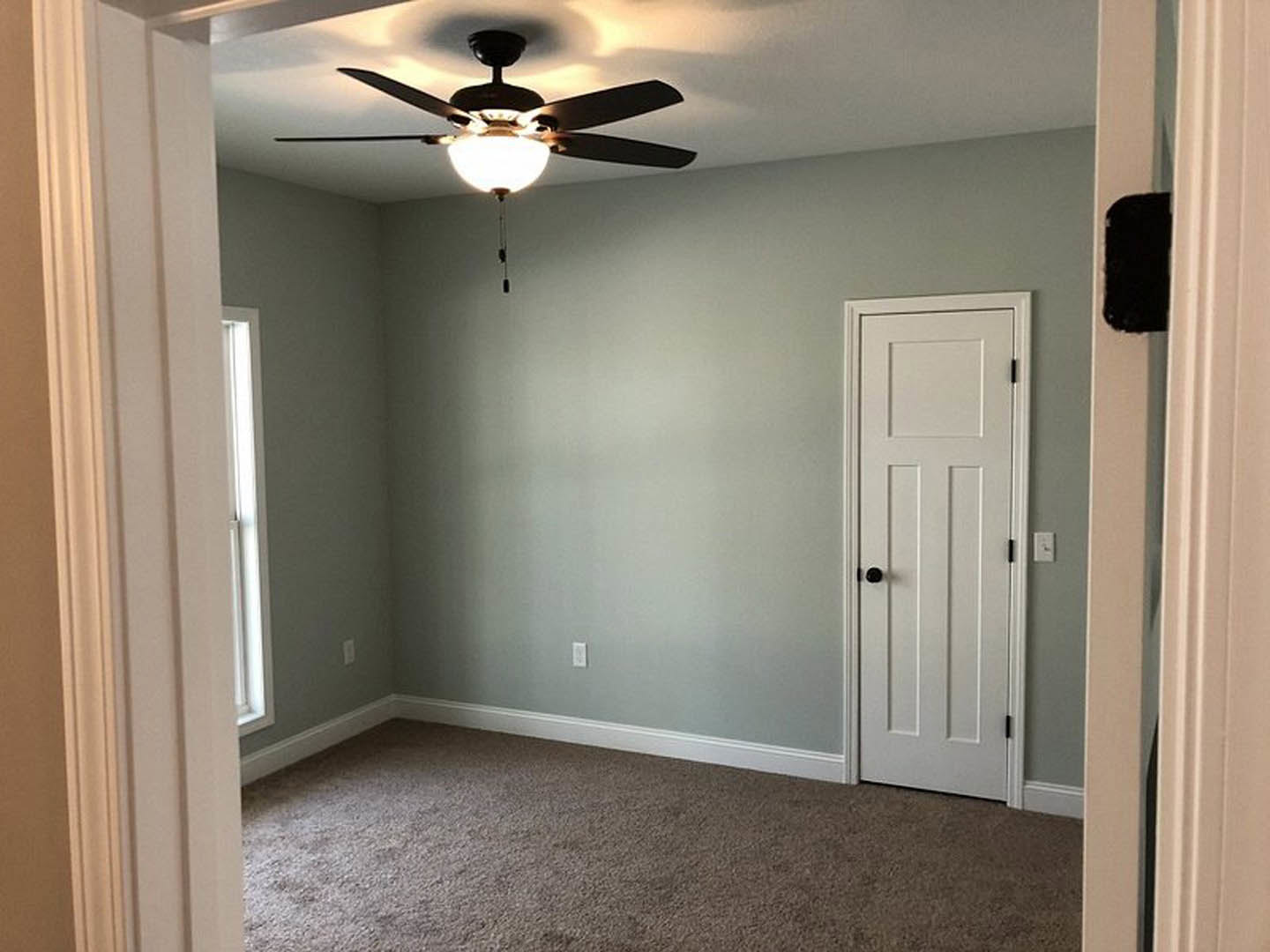 Carpeted room with white paneled door featuring a window, ceiling fan with light fixture, plaster walls, and neutral finishes.