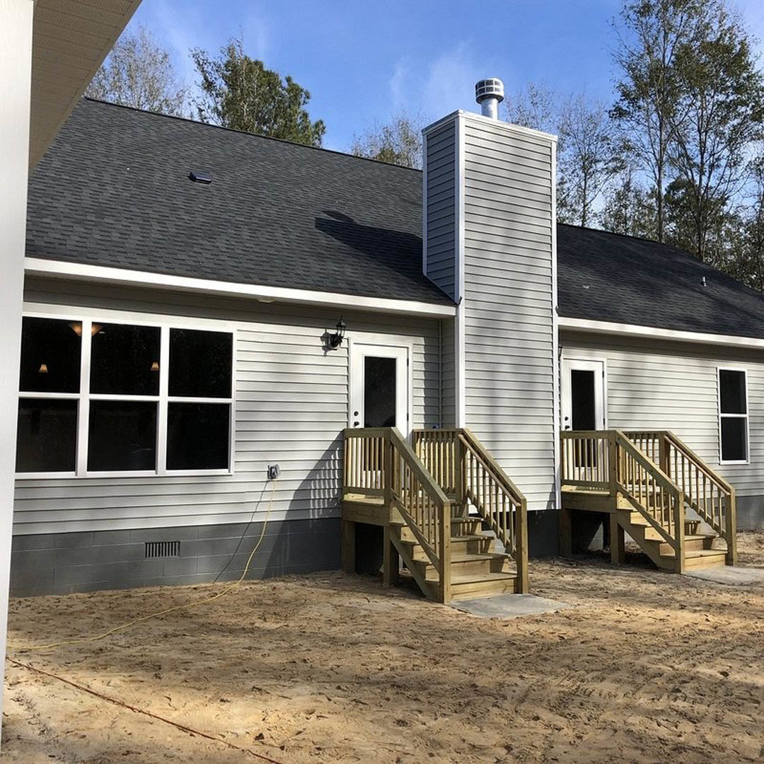 Wooden exterior stairs leading to a porch, sandy ground in front, large windows and tower detail on modern siding, surrounded by trees under blue sky